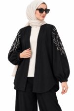 Cardigan long noir à motifs argentés sur les manches, vu de trois quarts sur une mannequin voilée tenant le bord du cardigan