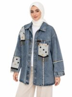Femme voilée légèrement tournée, les bras détendus, portant une veste en jean bleu délavé ornée d’étoiles noires et dorées